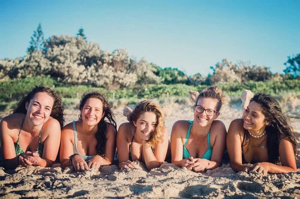 grupo de chicas en bikini estiradas y mirando a cámara sonrientes. Están estiradas encima de la arena y detrás hay la vegetación